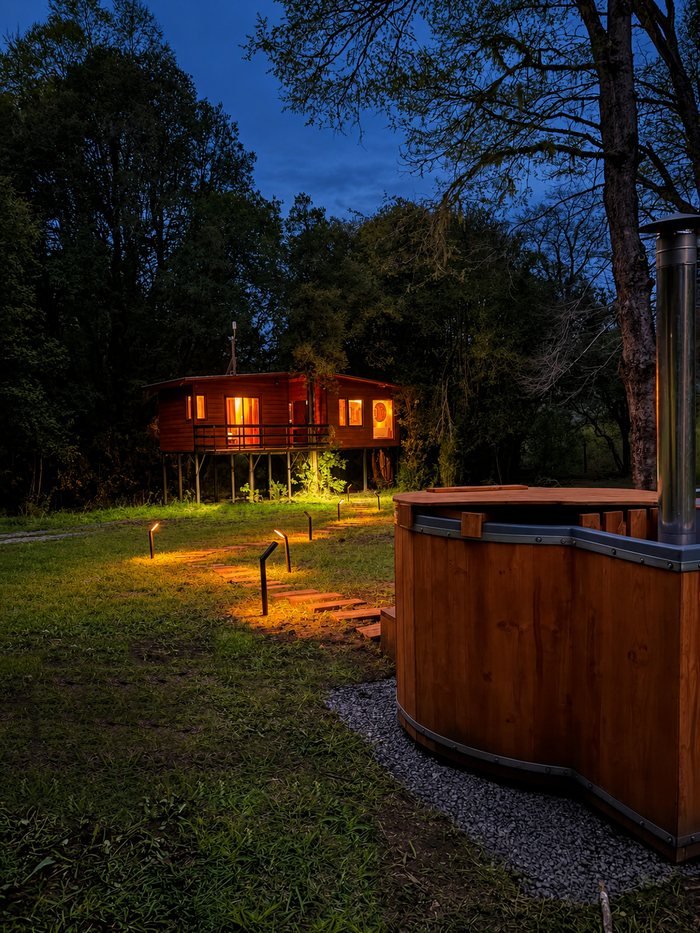 La cabaña iluminada al anochecer, con el sendero de luces y la tinaja de madera en primer plano